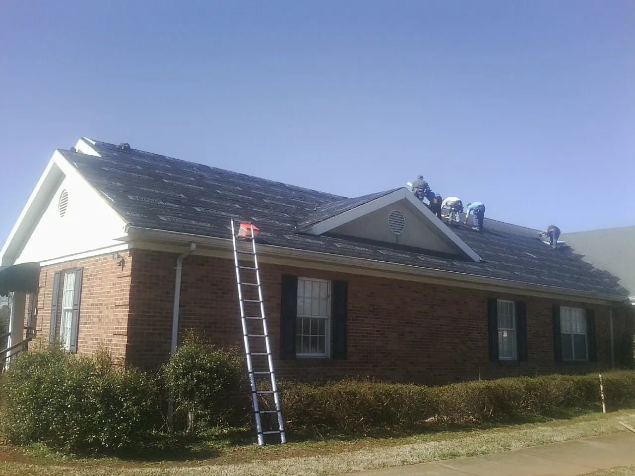 Crew tearing shingles off a brick ranch roof with a ladder set — Emergency Roof Repair work by our crew in Greens Fork