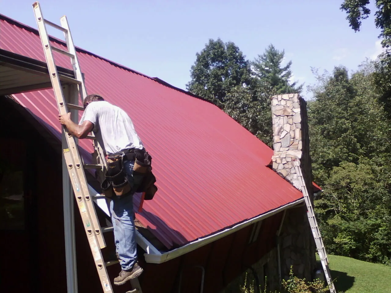 Roofer on a ladder beside a red metal roof and stone chimney — Soffit & Fascia Repair by Roofing Brothers in Greens Fork
