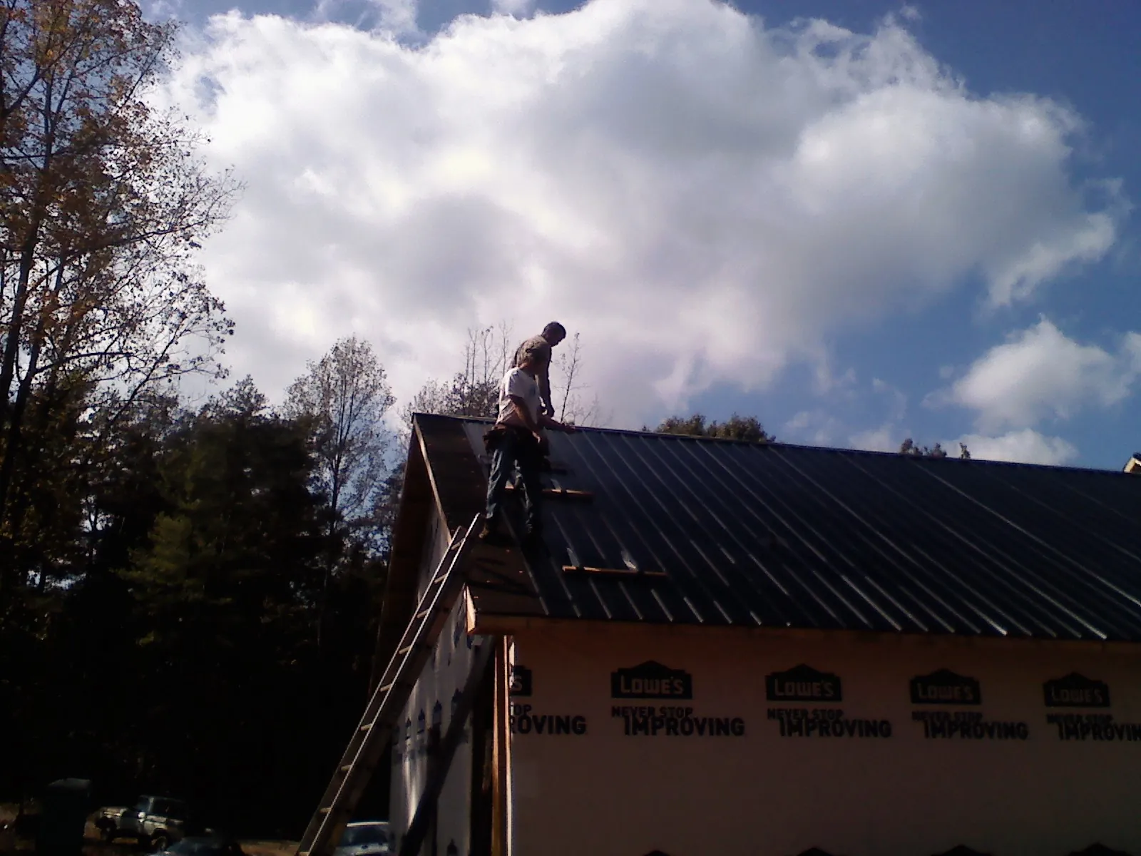 Roofer fastening dark metal panels high against a cloudy sky, from a Roof Inspection job in Greens Fork